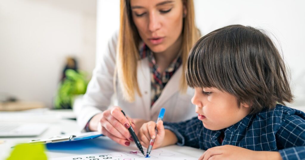 Niño concentrado aprendiendo a escribir números con ayuda de una maestra o terapeuta, usando marcadores de colores.
