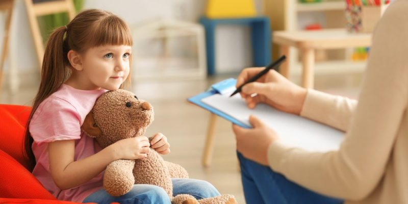 Niña con su oso de peluche sentada en un bean bag, mirando pensativamente mientras un terapeuta o psicólogo infantil toma notas.
