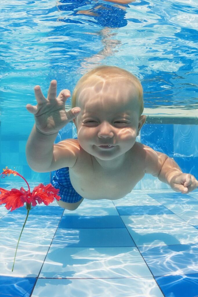 Bebé rubio sonriente nadando bajo el agua en una piscina con una flor roja.