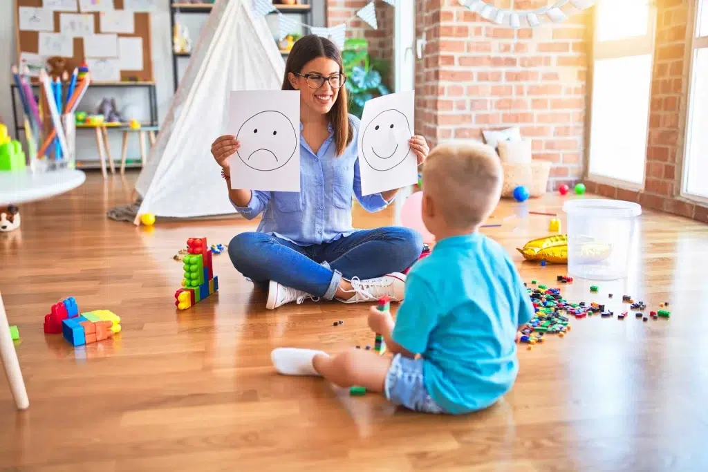 Terapeuta o maestra enseñando emociones (tristeza y felicidad) a un niño pequeño usando tarjetas con caras en un entorno de juego.