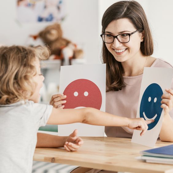 Terapeuta sonriente enseñando a un niño a reconocer emociones con tarjetas de caras felices y tristes.